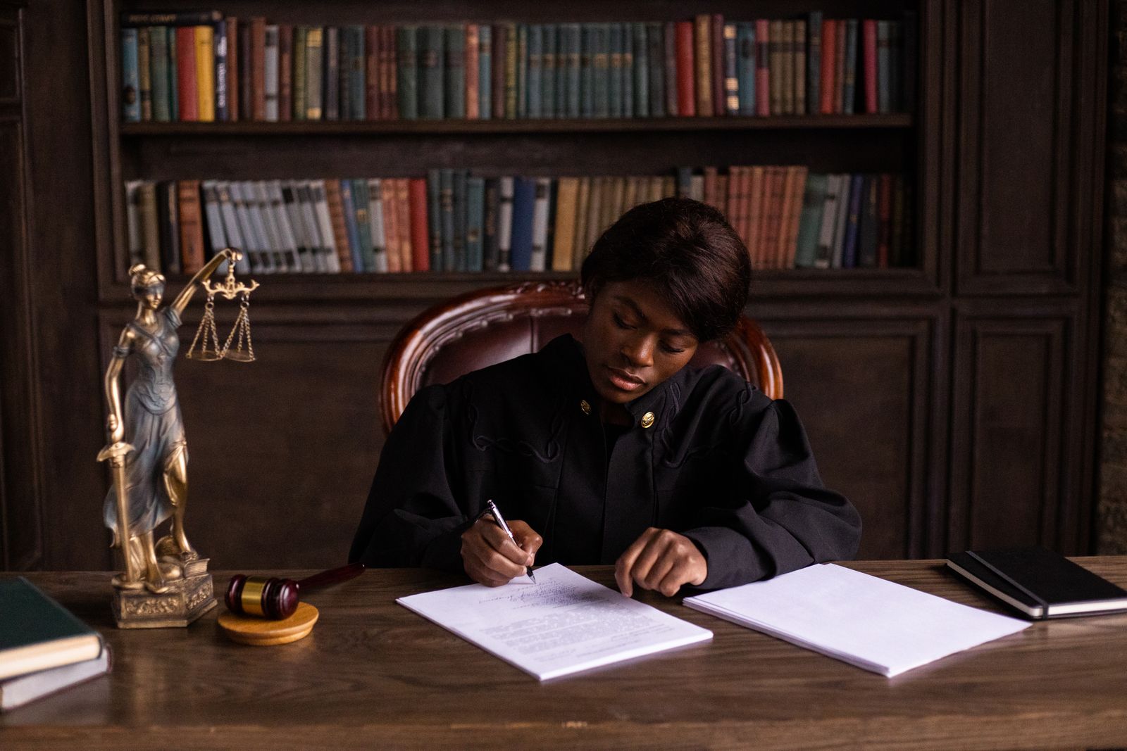 An advocate drafting by hand at a library desk, a figure of Justice at her side.